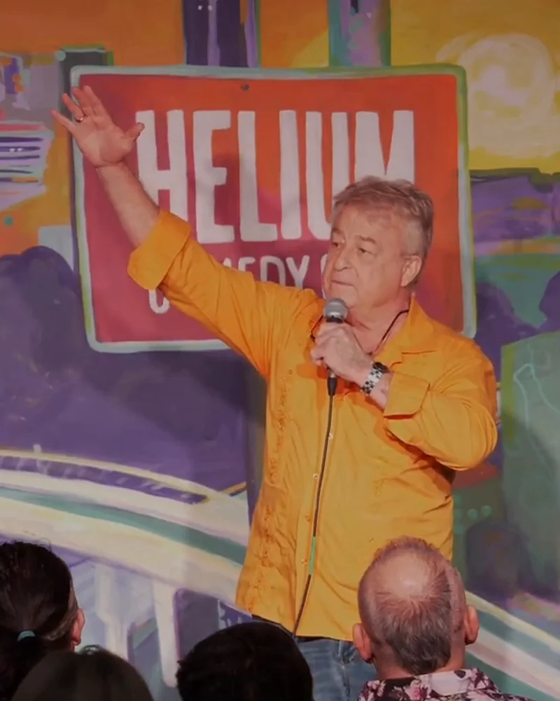 Uncle G in an orange shirt, arm raised, at Helium Comedy Club